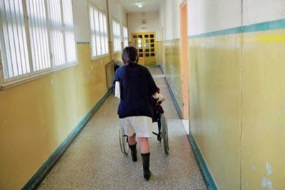 γυναίκα σπρώχνει ασθενή σε καροτσάκι μέσα σε ένα διάδρομο ψυχιατρείου